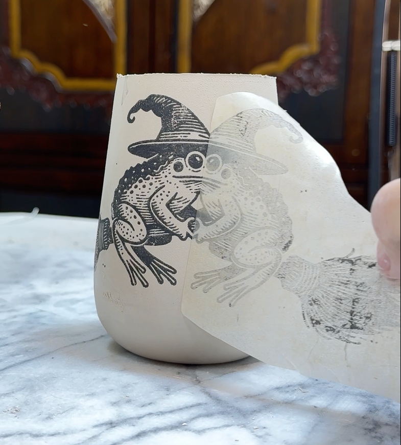 Frog design being applied to a mug using underglaze transfer paper